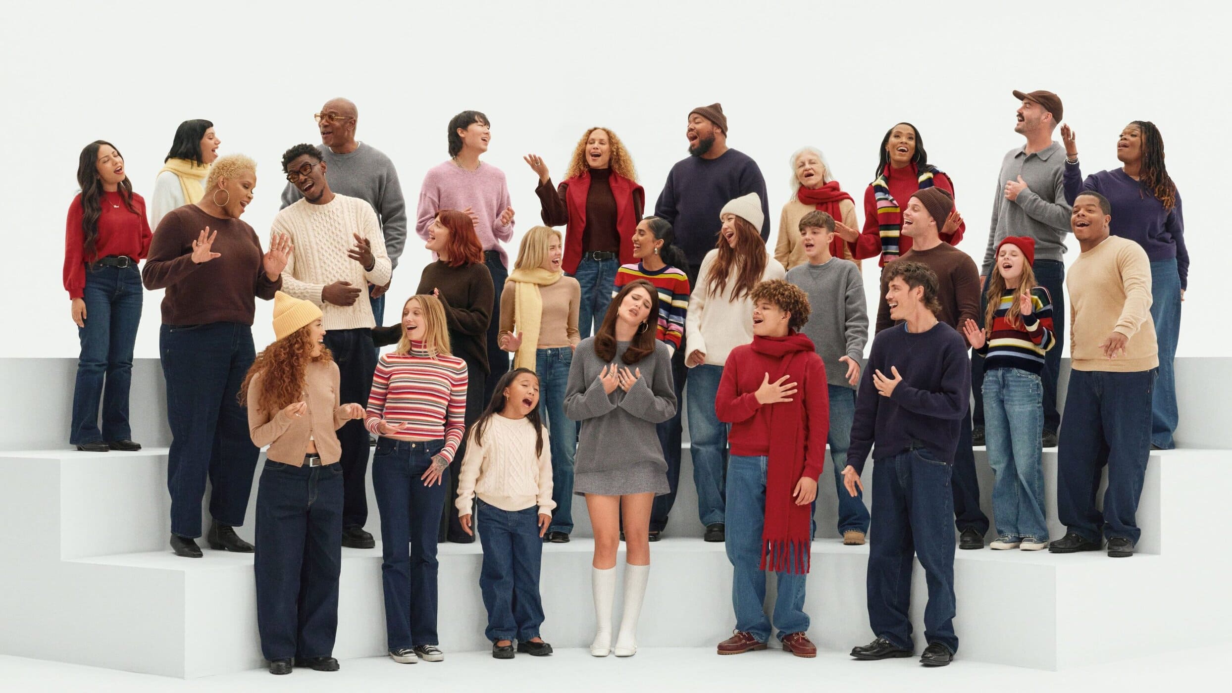 A diverse group of 25+ adults and children standing on white steps, singing in an indoor studio setting wearing sweaters, jeans, and scarves.