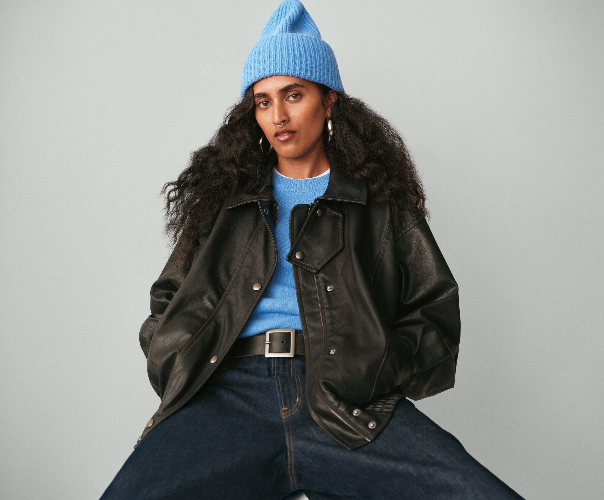Portrait of woman with dark hair in blue knit hat, light blue sweater, black jacket, and blue jeans posing against light gray backdrop.
