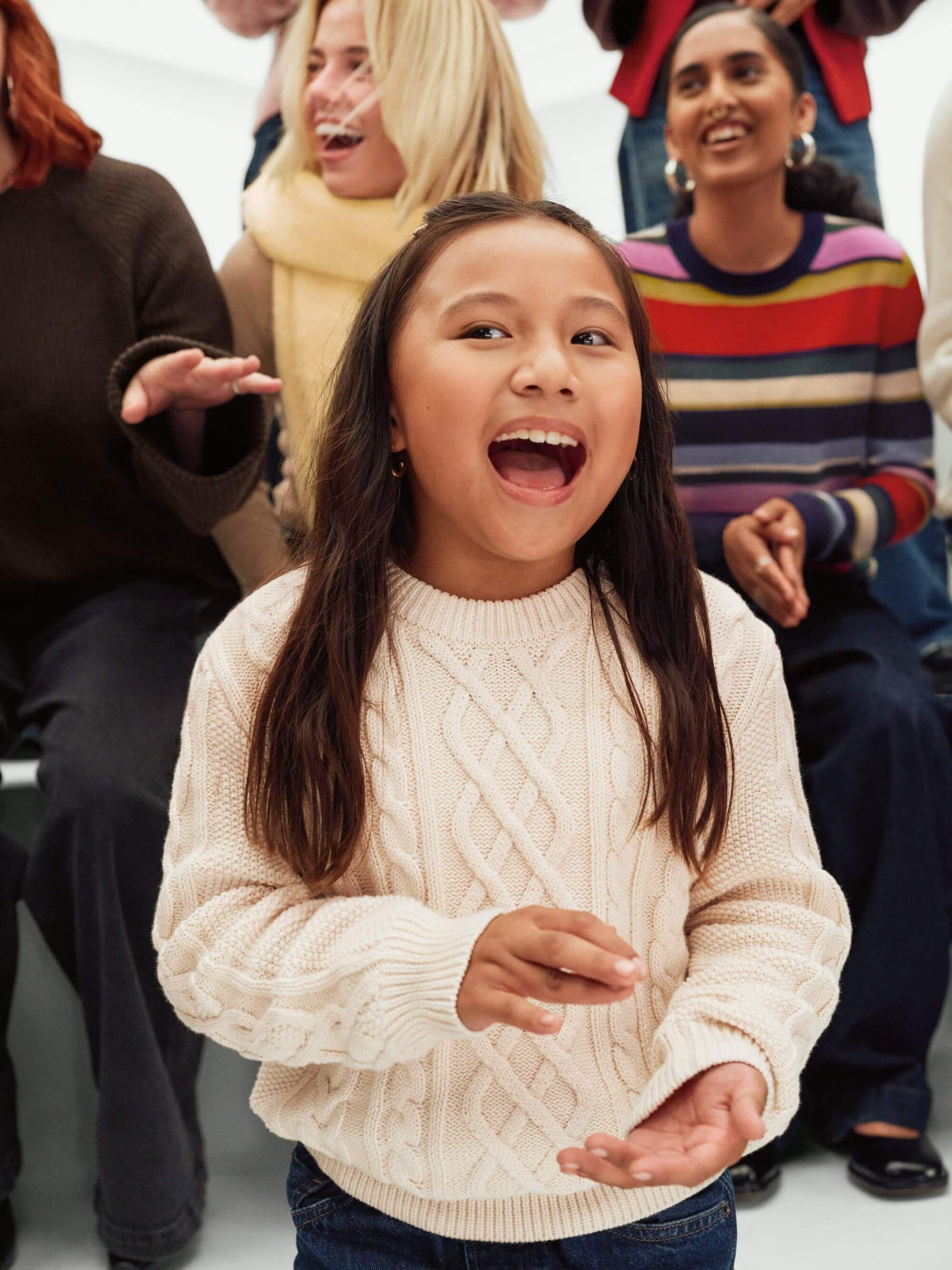 Portrait of smiling child wearing cream cable-knit sweater with a diverse group of adults behind her in casual colorful clothing.