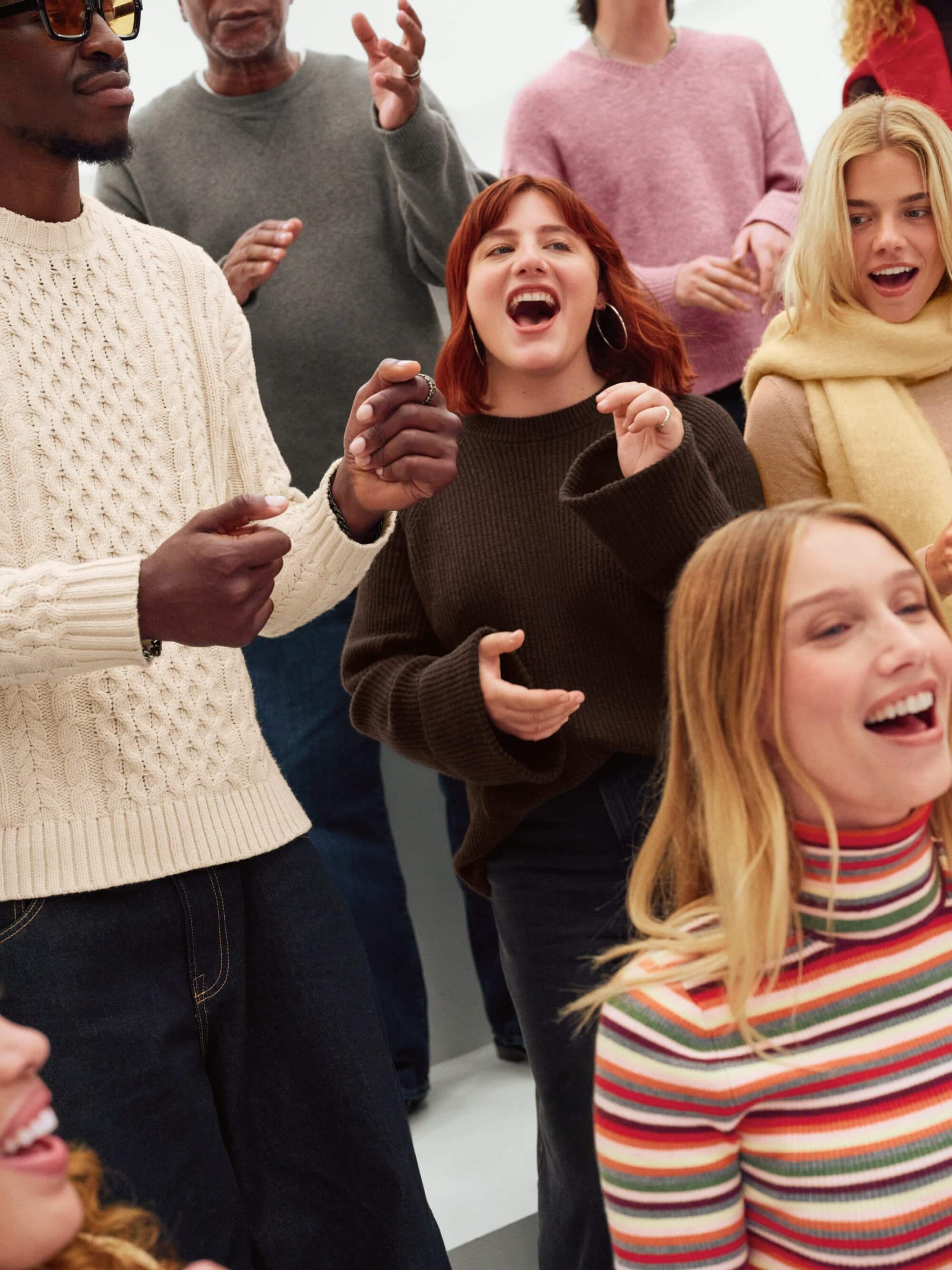 Group of diverse adults singing indoors, wearing casual sweaters and tops. Several people have their mouths open in song; indoor environment.