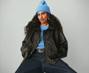 Model wearing a blue beanie, black leather jacket, and blue sweater against a grey backdrop for Gap Holiday 2025 campaign.