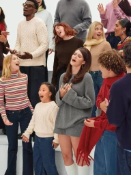 Group of diverse people in winter apparel singing and gesturing against a bright white background for Gap Holiday 2025 campaign.