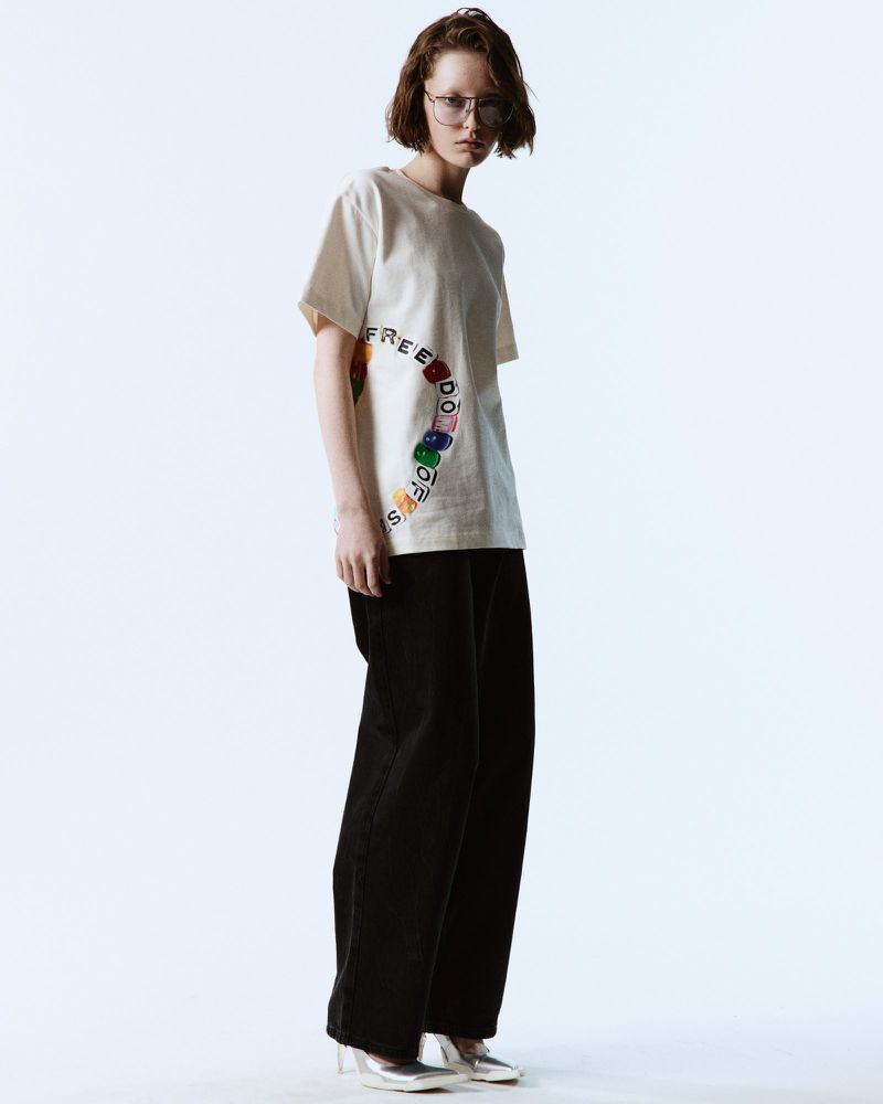 Adult woman in casual off-white tee with rainbow lettering and wide-leg black pants, silver shoes standing in a studio environment.
