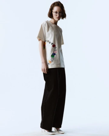 Adult woman in casual off-white tee with rainbow lettering and wide-leg black pants, silver shoes standing in a studio environment.
