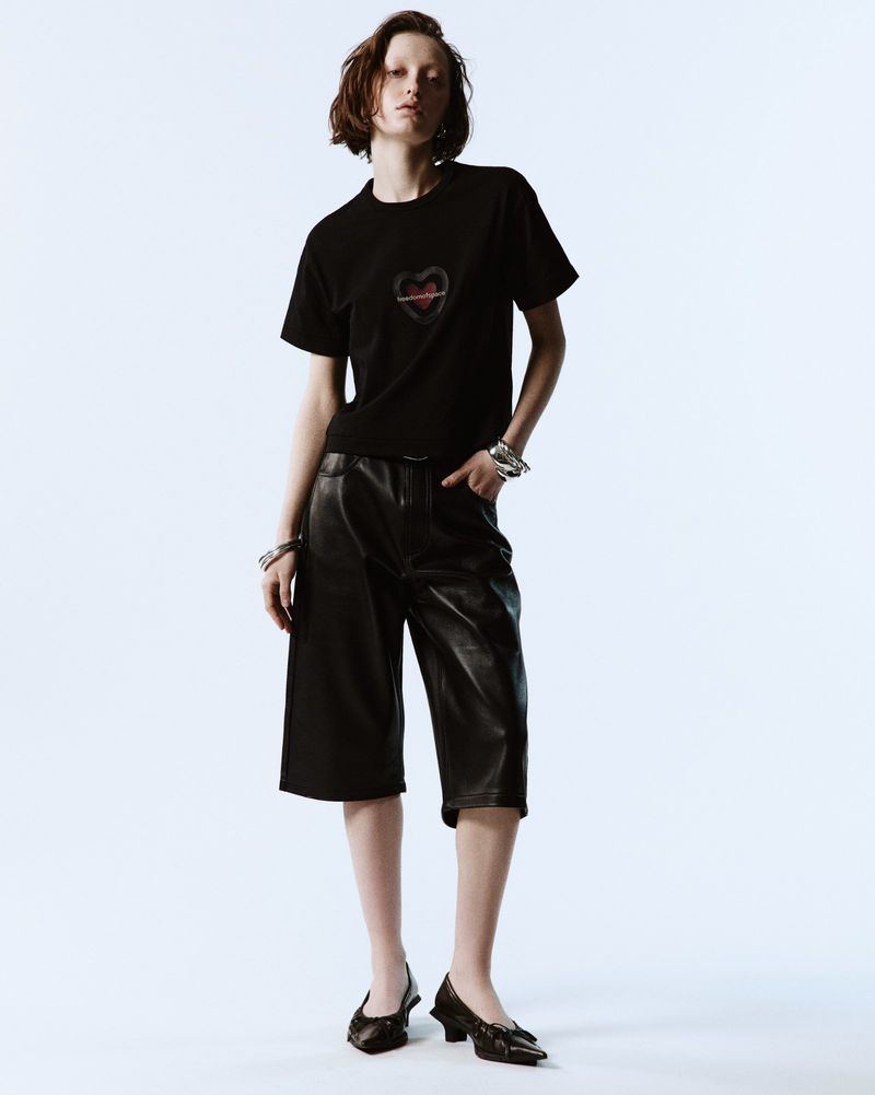Fashion shot of a woman with short hair wearing a black T-shirt, black leather culottes, and high heels against a white backdrop.
