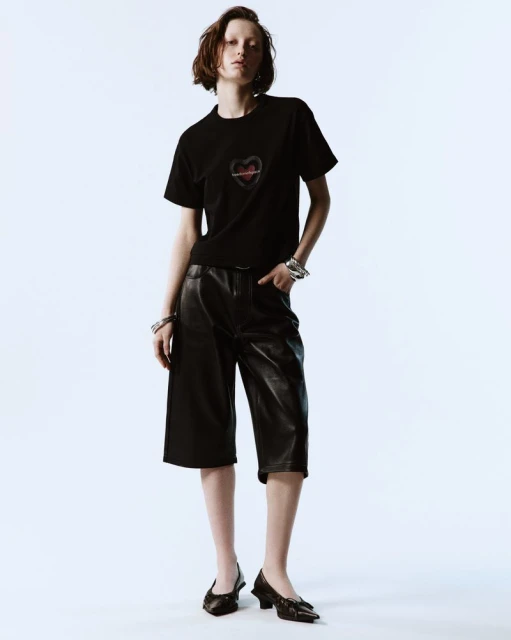 Fashion shot of a woman with short hair wearing a black T-shirt, black leather culottes, and high heels against a white backdrop.
