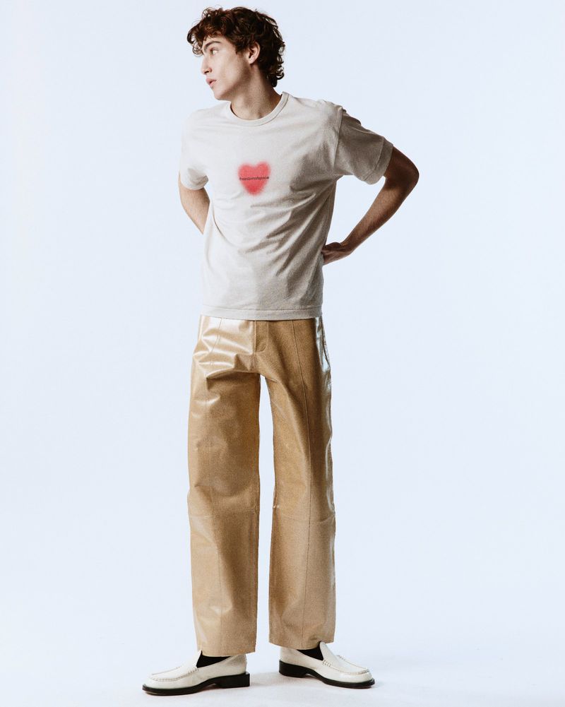 Full-body shot of a young adult male in a beige t-shirt with a heart design, tan pants, and white loafers posing in a brightly lit photo studio.