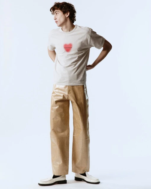 Full-body shot of a young adult male in a beige t-shirt with a heart design, tan pants, and white loafers posing in a brightly lit photo studio.