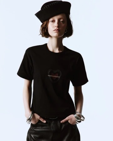 Portrait of a young woman wearing a black t-shirt with a heart graphic, paired with leather pants and silver bracelets against a white backdrop.