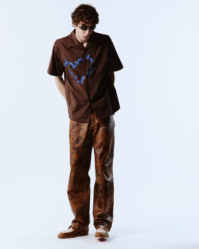 A young adult man poses in a brown collared shirt with a blue graphic design, paired with brown pants and shoes, against a white backdrop.