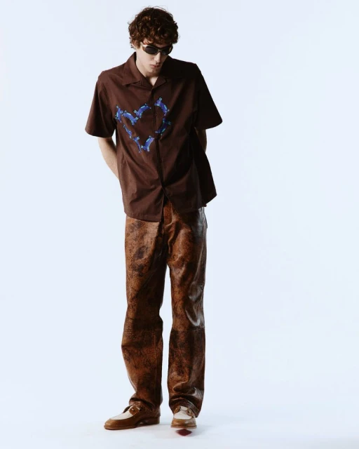 A young adult man poses in a brown collared shirt with a blue graphic design, paired with brown pants and shoes, against a white backdrop.