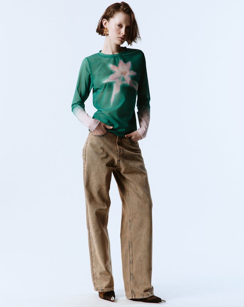 A young woman poses in a fashion shot wearing a green floral-print long-sleeved top and brown wide-leg jeans against a white backdrop.