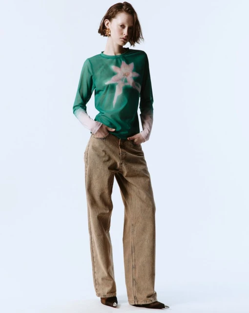 A young woman poses in a fashion shot wearing a green floral-print long-sleeved top and brown wide-leg jeans against a white backdrop.