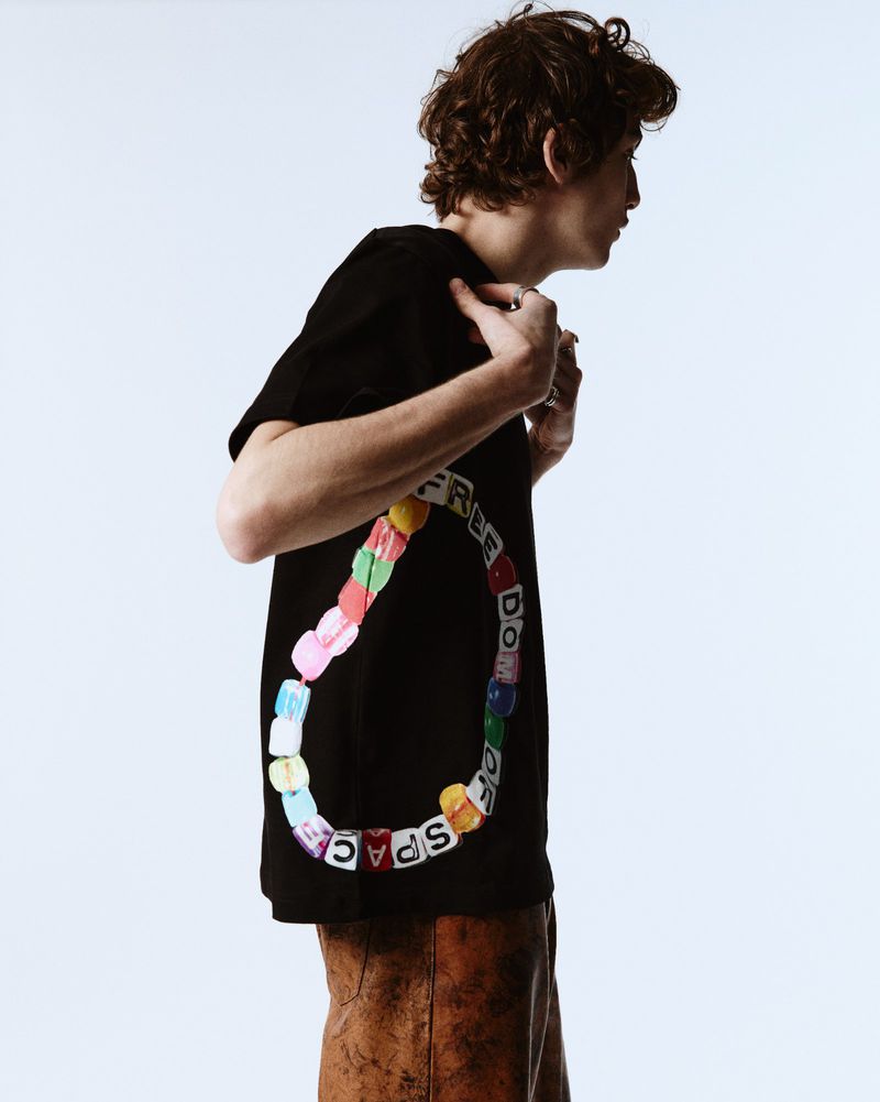 Young adult poses in black t-shirt with colorful graphic design against bright white backdrop. Fashion-forward style, curly hair.