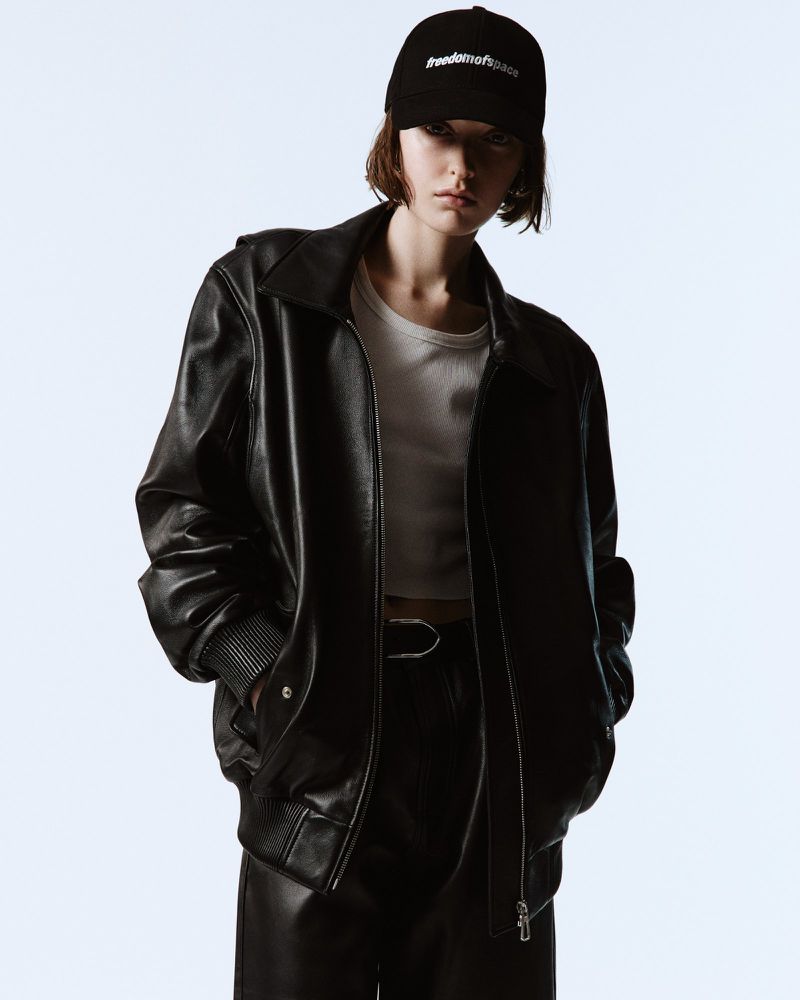 A young adult woman in a black leather jacket, white crop top, and cap poses in a studio with a light blue background in a fashion shot.