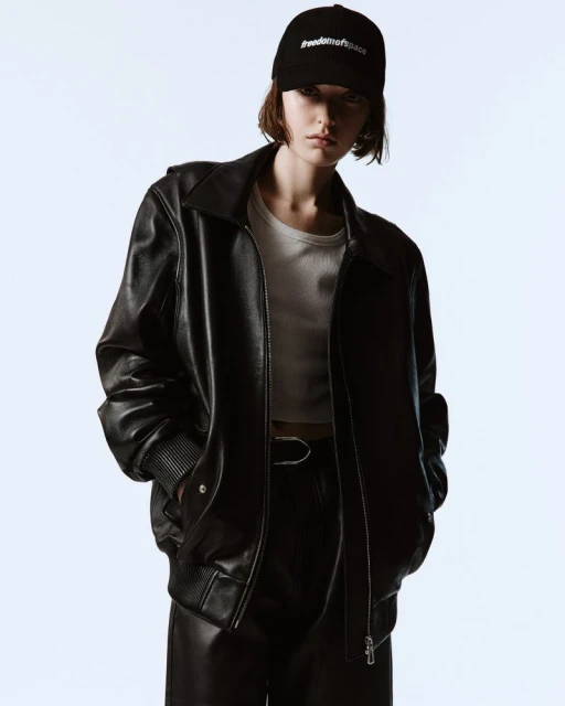 A young adult woman in a black leather jacket, white crop top, and cap poses in a studio with a light blue background in a fashion shot.