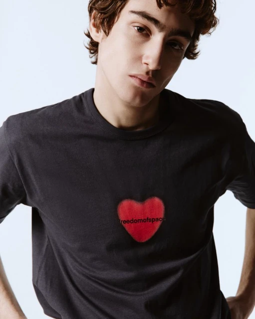 Close-up of a young man wearing a dark t-shirt with a red heart graphic and the text 'freedomofspace'. Light background, soft lighting.