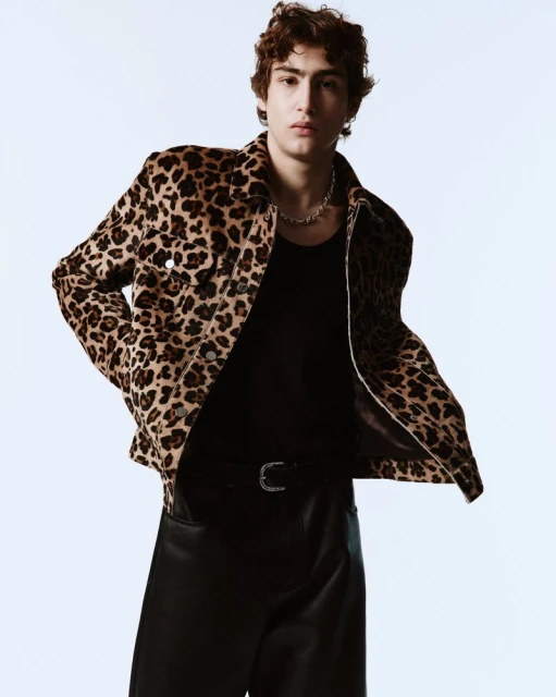 A young adult male with curly hair poses in a studio wearing a leopard print jacket, black shirt, black pants, and a silver chain.