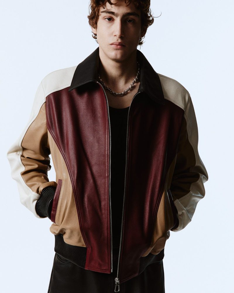 A young man with curly hair wearing a burgundy, brown, and white leather bomber jacket and a silver chain on a white background.