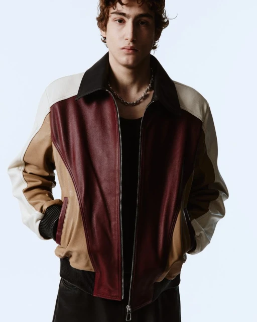 A young man with curly hair wearing a burgundy, brown, and white leather bomber jacket and a silver chain on a white background.