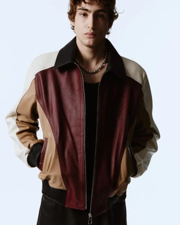 A young man with curly hair wearing a burgundy, brown, and white leather bomber jacket and a silver chain on a white background.