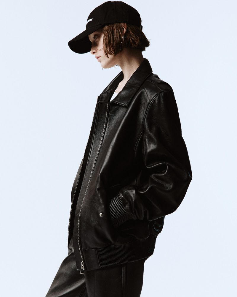 Model wearing a black leather jacket and black cap against a plain light background