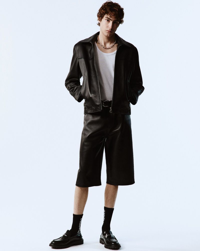 Young man standing against a white backdrop wearing a black leather jacket, shorts, white t-shirt, and black loafers.