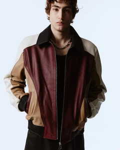 Young man wearing a color-blocked leather jacket and silver chain necklace against a plain white studio background.