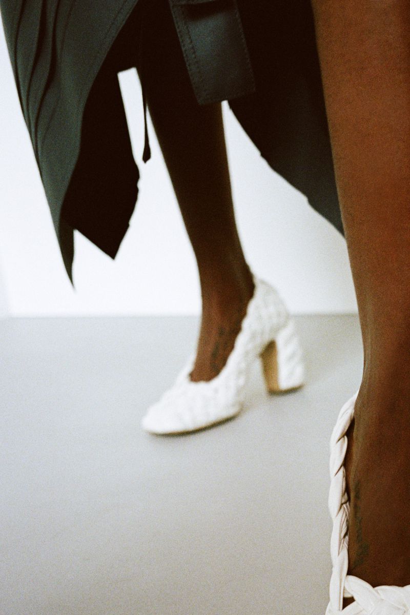 Close-up shot of an adult female wearing elegant white braided-texture high-heeled shoes and a dark blue dress in a white studio.