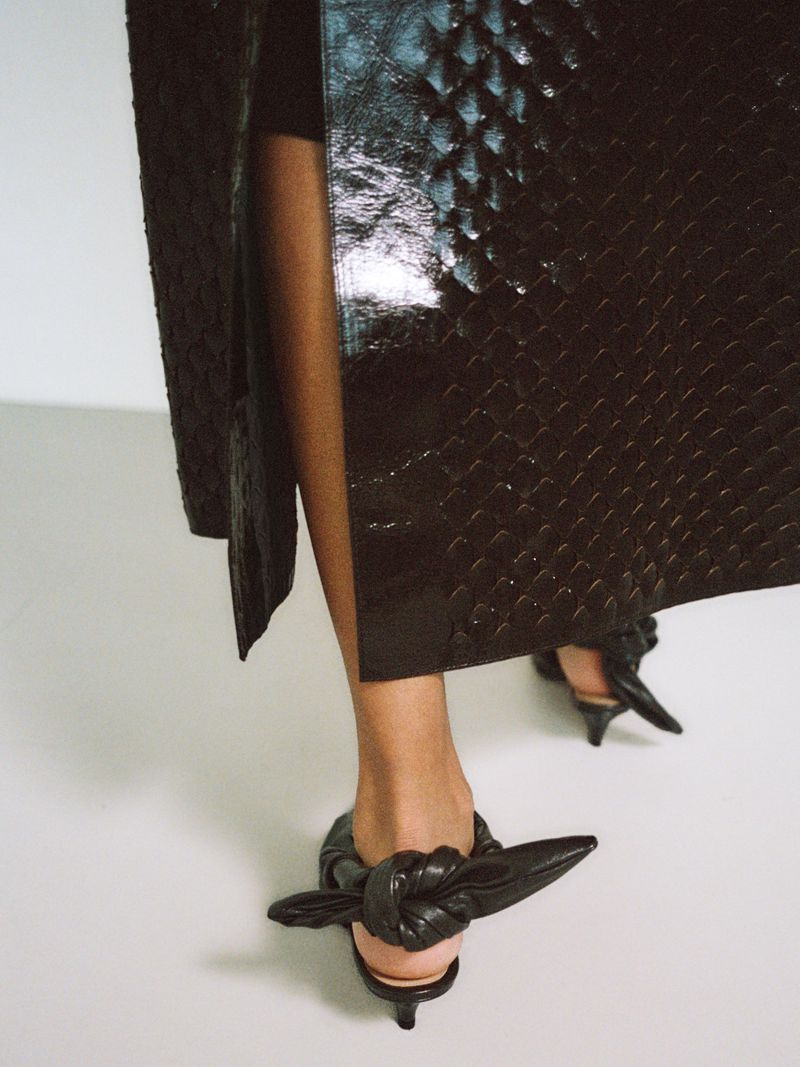 Woman in textured black skirt and high-heeled shoes, indoors on a white surface, fashion-forward footwear and apparel, studio shot.