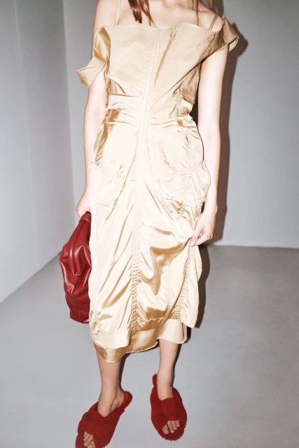 A woman in an off-the-shoulder beige dress holds a red bag, with red fuzzy sandals, posing in a minimalist interior setting with natural light.