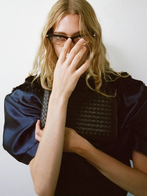 A blonde adult female wearing eyeglasses holds a black woven leather clutch bag in front of a white wall background.