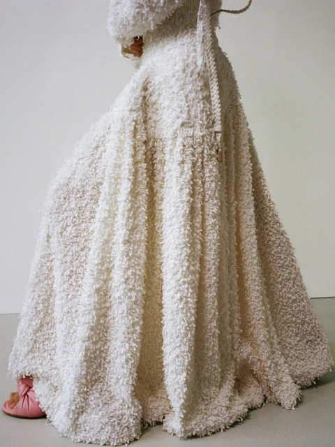 Person wearing a large, textured white robe and head covering, with light pink slippers visible underneath the hem, against a light background.