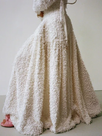Person wearing a large, textured white robe and head covering, with light pink slippers visible underneath the hem, against a light background.
