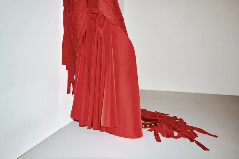 Elegant flowing red gown with detailed fabric and ribbons is styled against a clean white backdrop in an indoor fashion presentation.