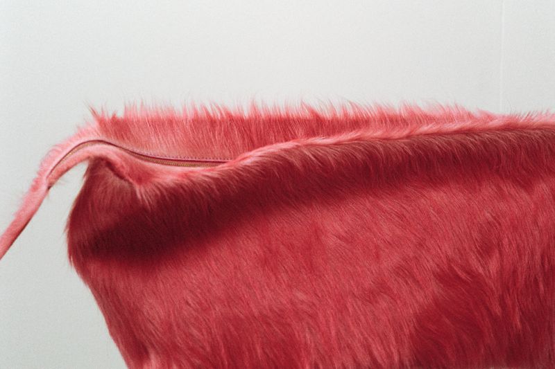 Close-up of a textured, reddish-pink handbag with a faux fur-like finish, set against a plain white backdrop in a studio setting.