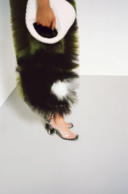 Close-up of a person wearing clear heels and a textured dress, carrying a white bag in a minimalist studio setting.