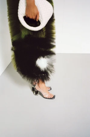 Close-up of a person wearing clear heels and a textured dress, carrying a white bag in a minimalist studio setting.