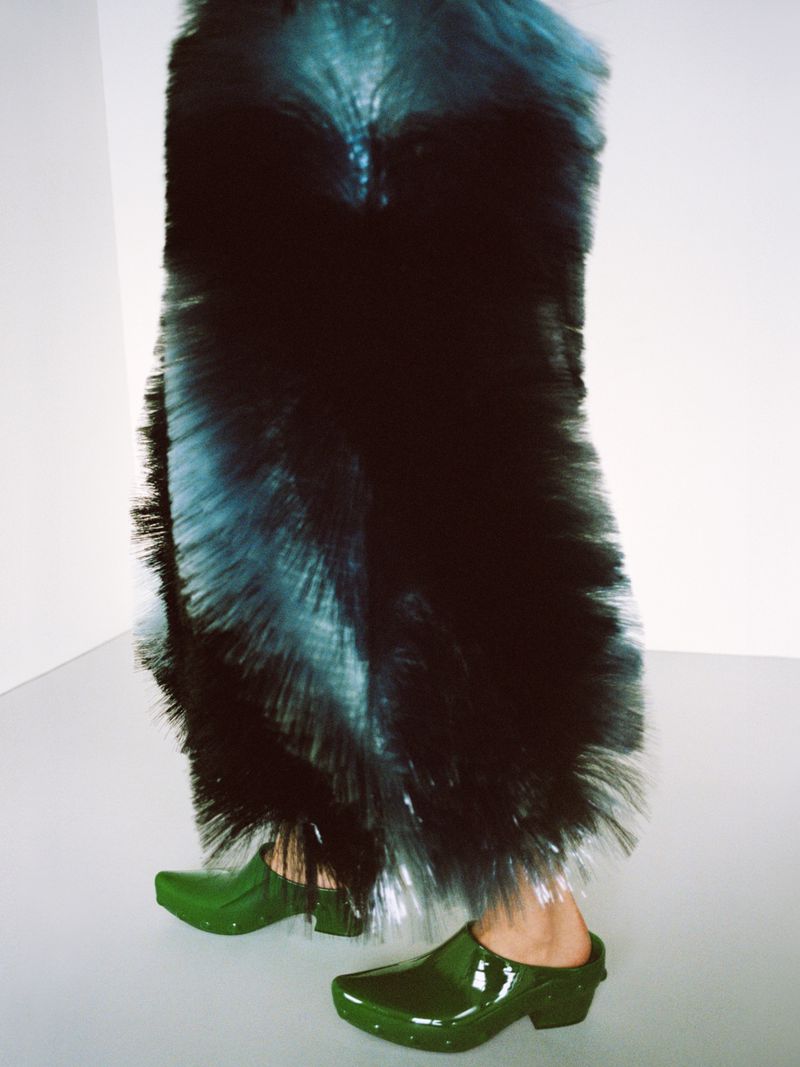 Woman in a large blue and black bristled skirt, matched with shiny green clogs, standing in a stark white room.