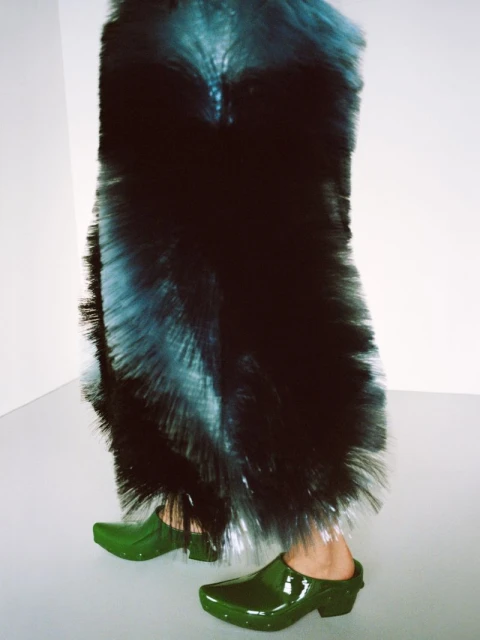 Woman in a large blue and black bristled skirt, matched with shiny green clogs, standing in a stark white room.
