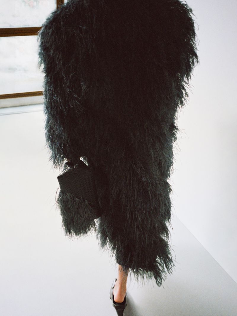 Fashionable woman in a black, feathered coat and heels holding a small purse in a light-filled, modern indoor setting.