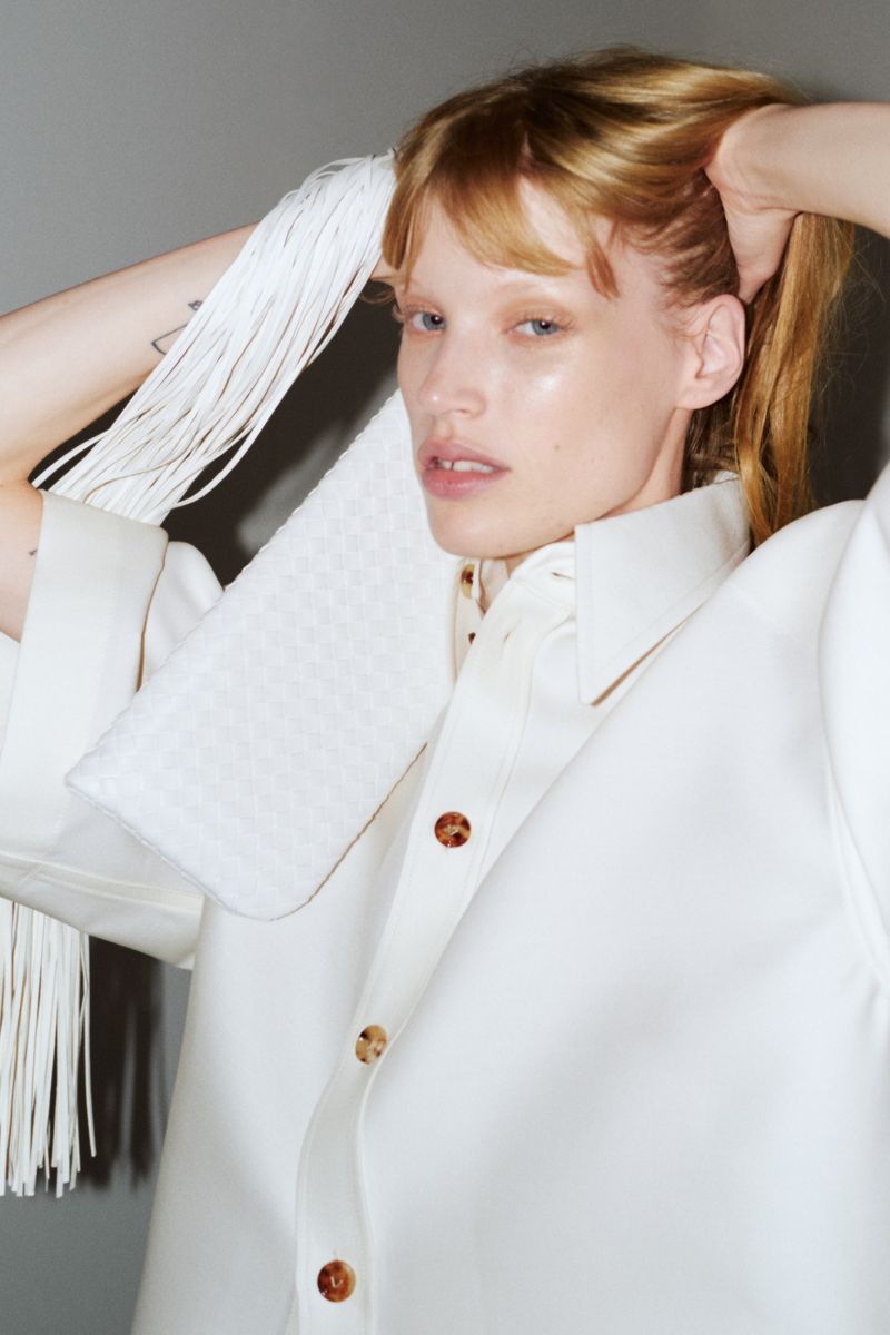 Model wearing a white shirt holding a white fringed bag against a grey background