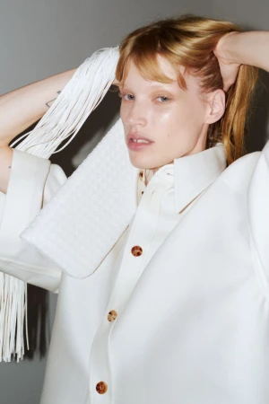 Model wearing a white shirt holding a white fringed bag against a grey background