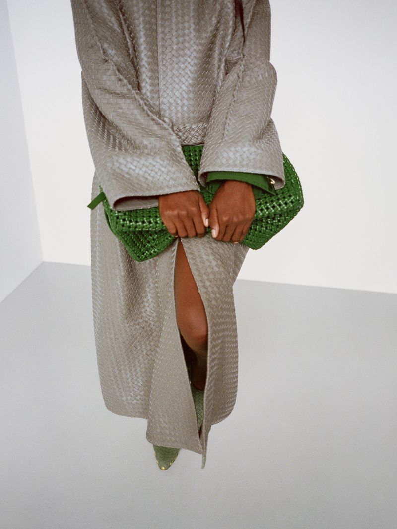 Fashionable woman wearing woven coat with split and green heels holding a woven green clutch bag in bright studio setting.