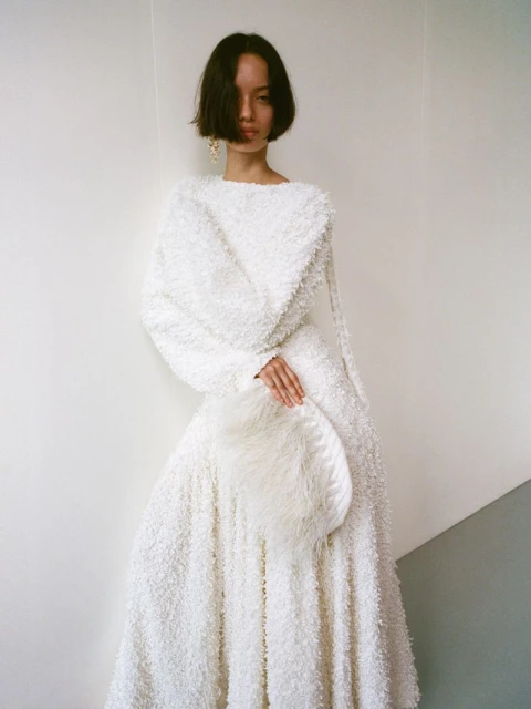 Elegant woman in a white textured dress holding a matching round handbag, posing against a neutral white wall indoors.