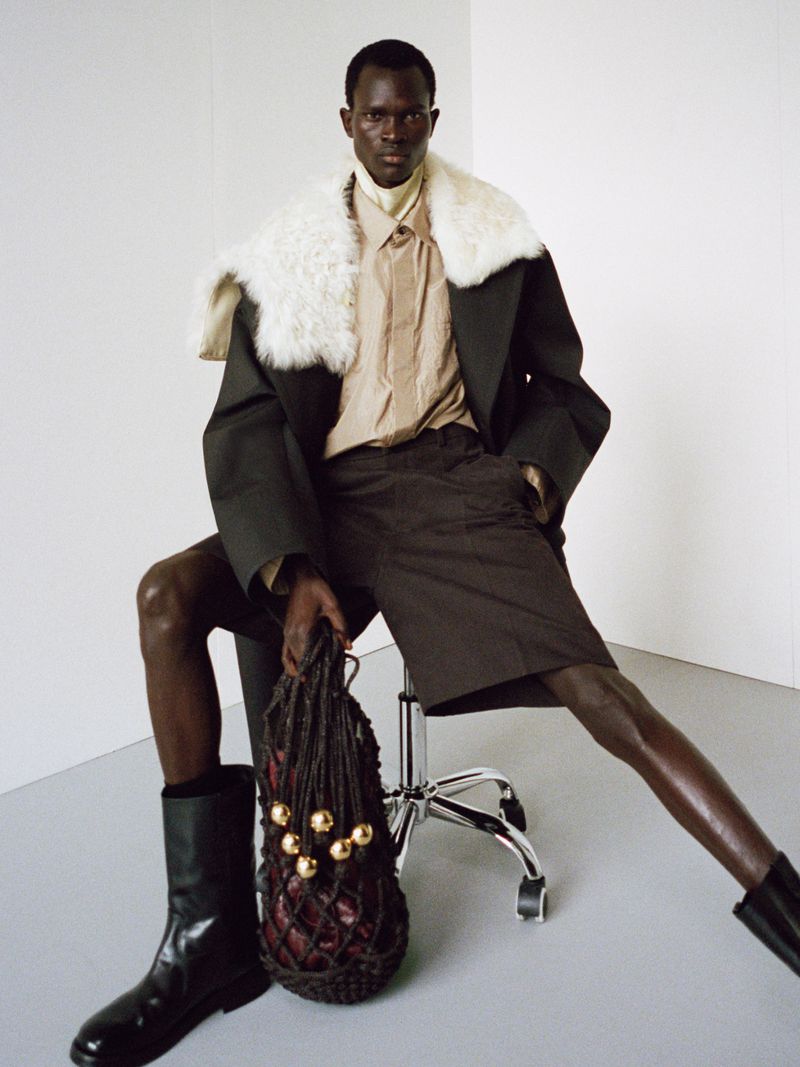 Man in black coat with fur collar sits on a rolling chair holding a net bag in a minimalist studio, captured in a fashion editorial shot.