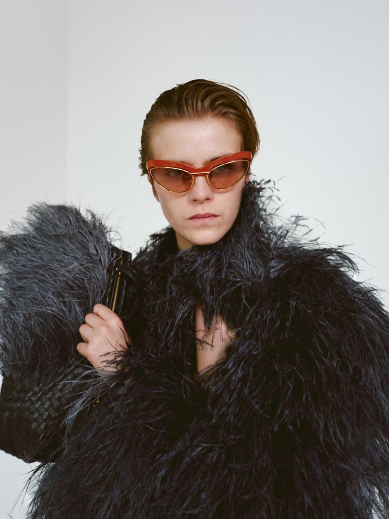 Portrait of a person with short hair wearing orange-framed sunglasses and a feathered garment against a plain white background.