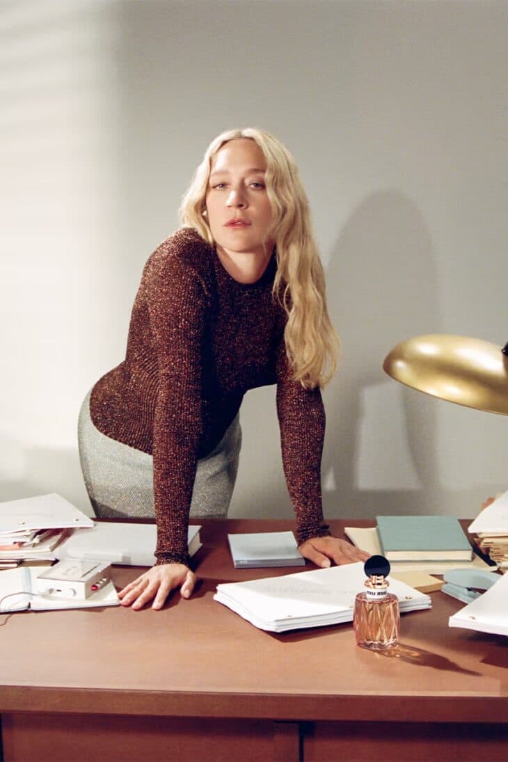Woman with blonde hair leans on a desk with paperwork, illuminated by a lamp, wearing a long-sleeved top and skirt in an indoor setting.