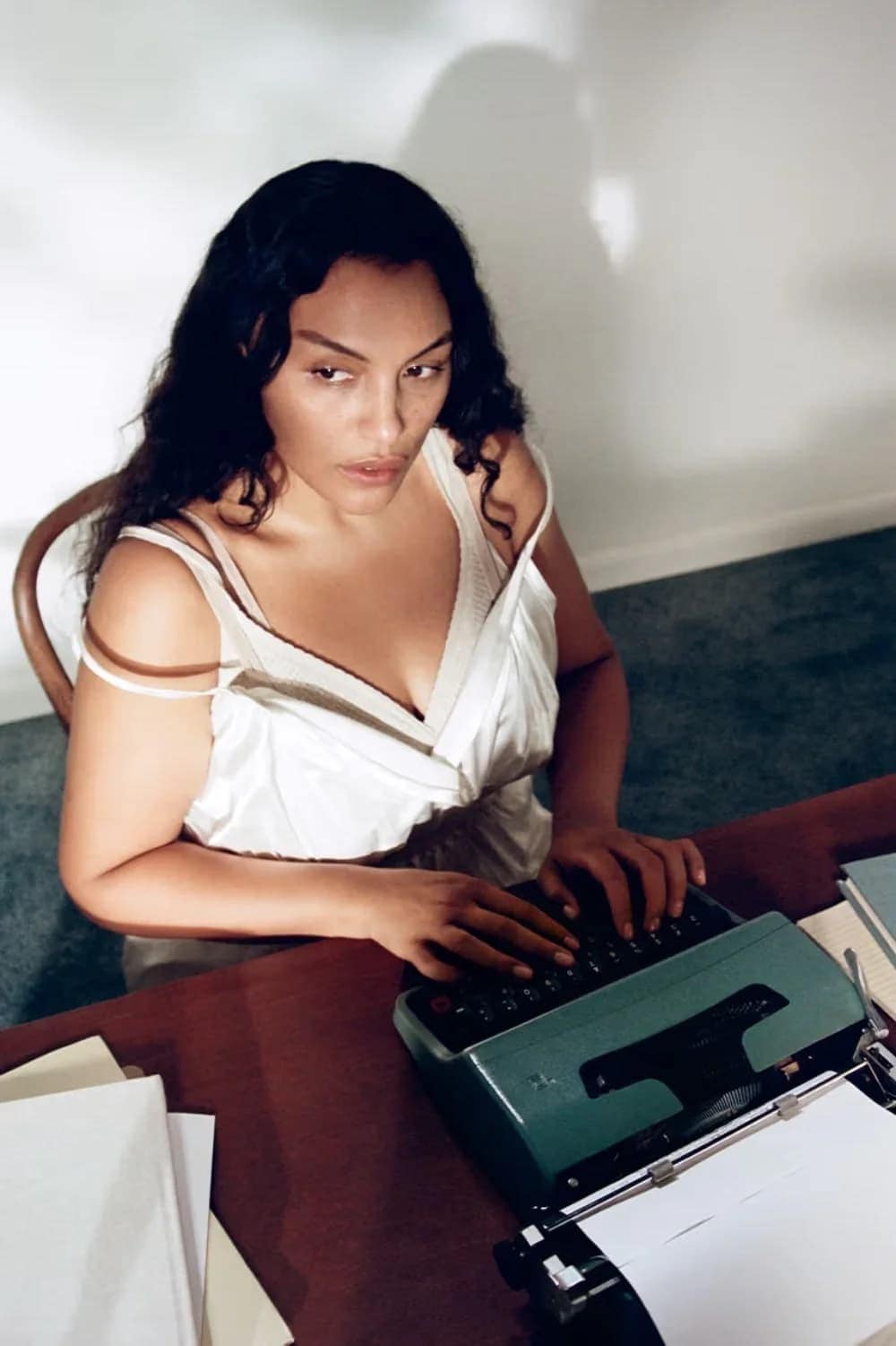 Woman types on a teal typewriter at a desk with stacks of paper in an indoor setting with soft lighting and a cream-colored wall.