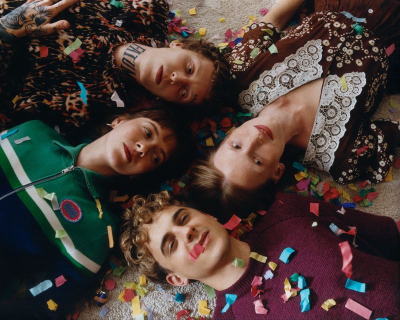 Four young adults, two women and two men, are laying head to head on a light-colored surface covered in colorful confetti pieces.